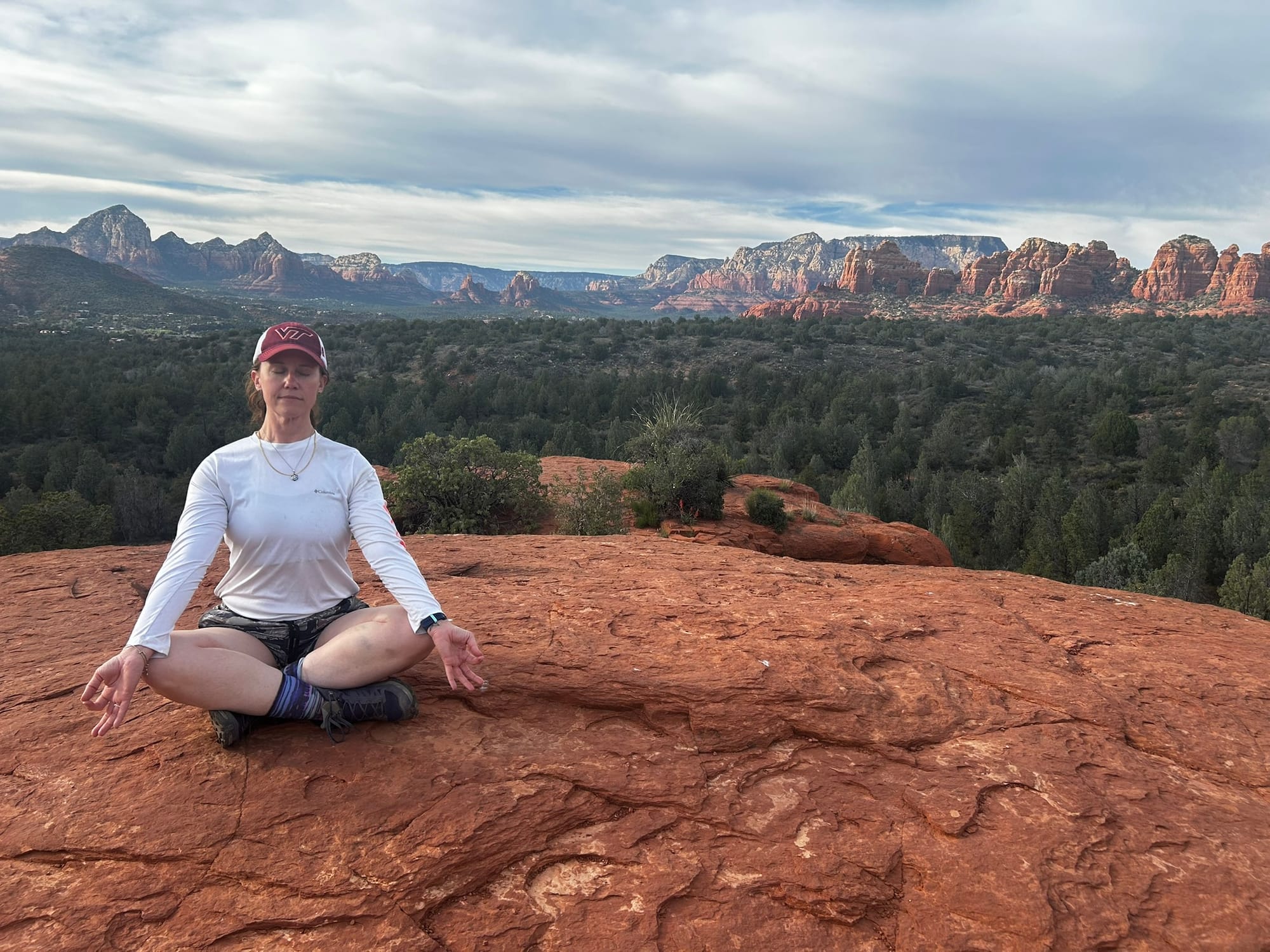 A woman sitting cross-legged meditating on red rocks with mountains and scrub pines in the background