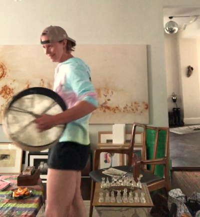 A person in a tie dyed shirt and backwards hat opening sacred space with a drum before performing shamanic work