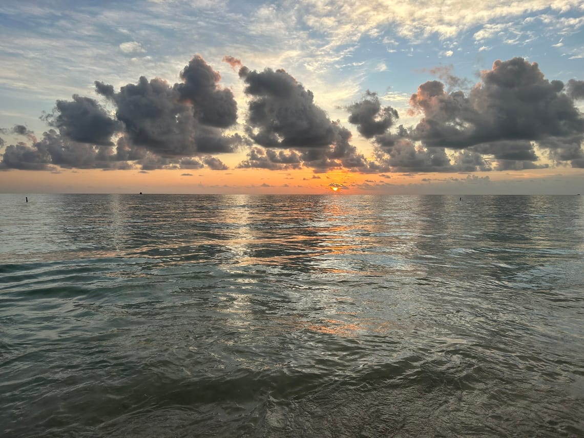 A blue gray body of water with gentle waves, an orange setting sun, and fluffy gray clouds